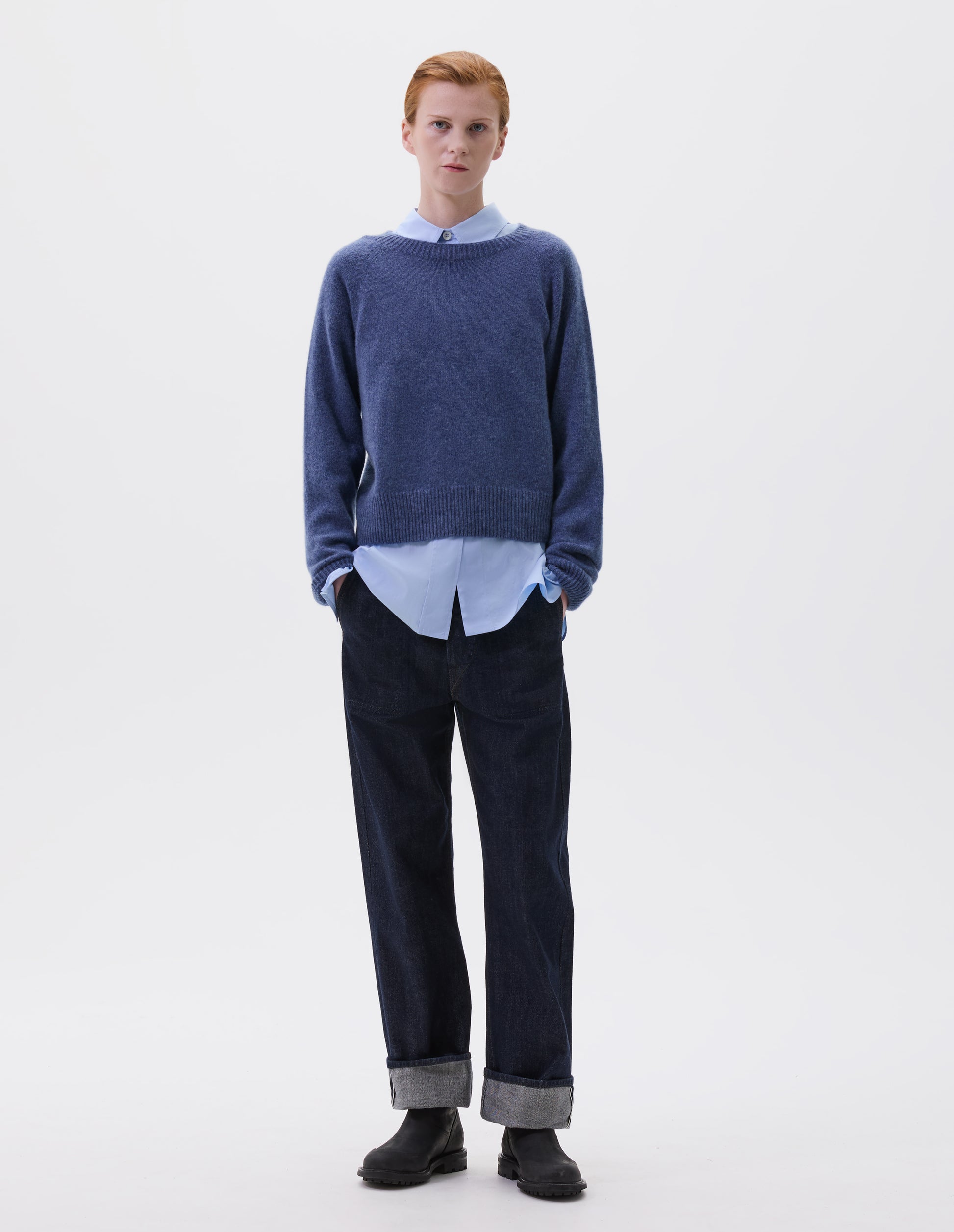 Full body image, front view of model wearing blue long sleeve cashmere jumper, with raglan sleeve and contrasting rib on neck, cuffs and hem. Styled over blue collared shirt, with indigo denim jeans and black leather boots.