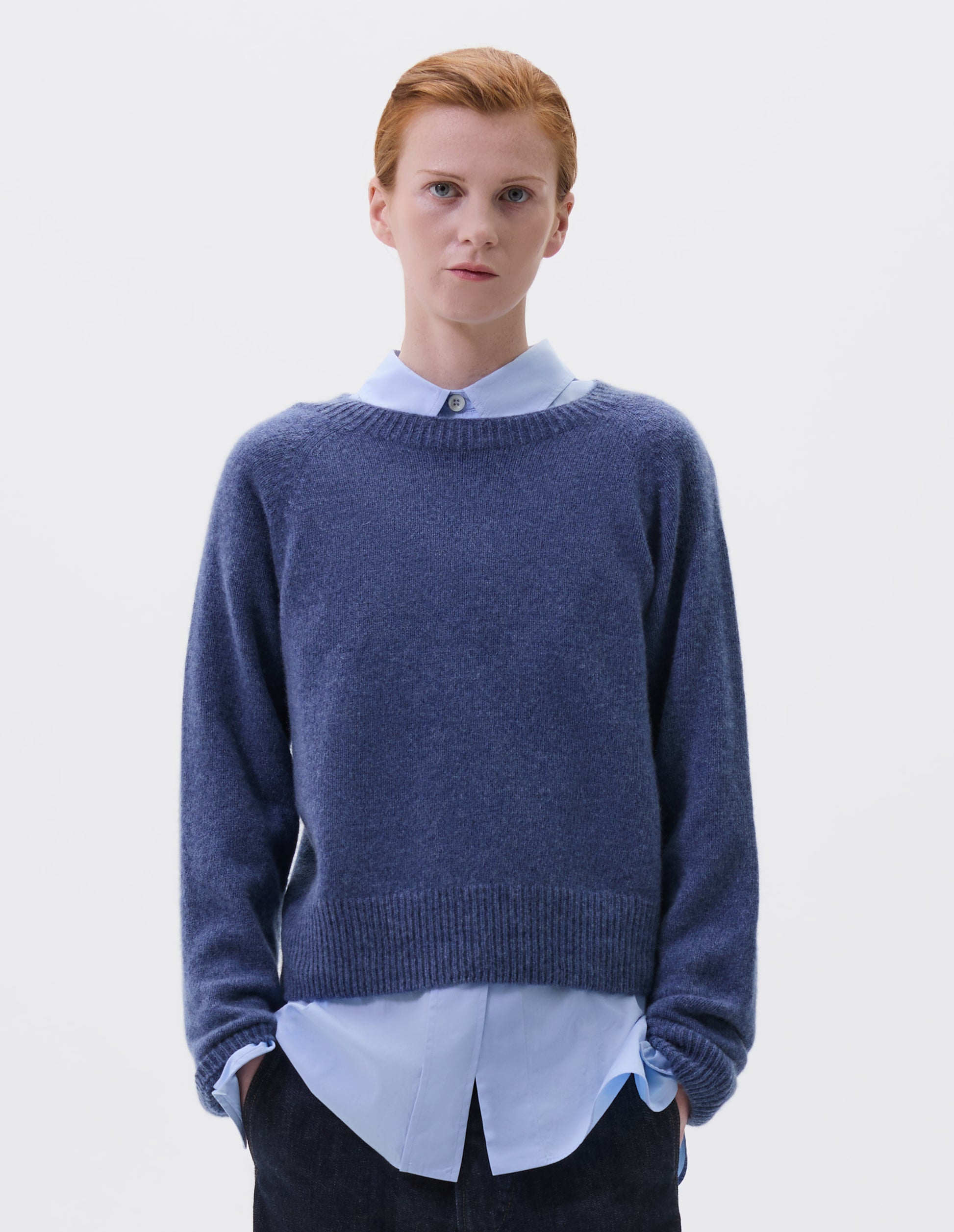 Cropped body image, front view of model wearing blue long sleeve cashmere jumper, with raglan sleeve and contrasting rib on neck, cuffs and hem. Styled over blue collared shirt, with indigo denim jeans.