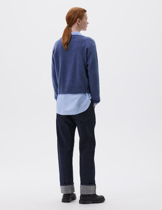 Full body image, back view of model wearing blue long sleeve cashmere jumper, with raglan sleeve and contrasting rib on neck, cuffs and hem. Styled over blue collared shirt, with indigo denim jeans and black leather boots.