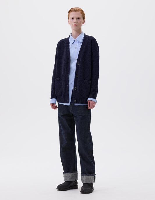 Full body image, front view of model wearing navy v-neck cardigan, with contrast rib on placket, cuffs and hem and two front pockets, finished with horn button closure. Styled over blue shirt, indigo blue jeans and black leather boots.