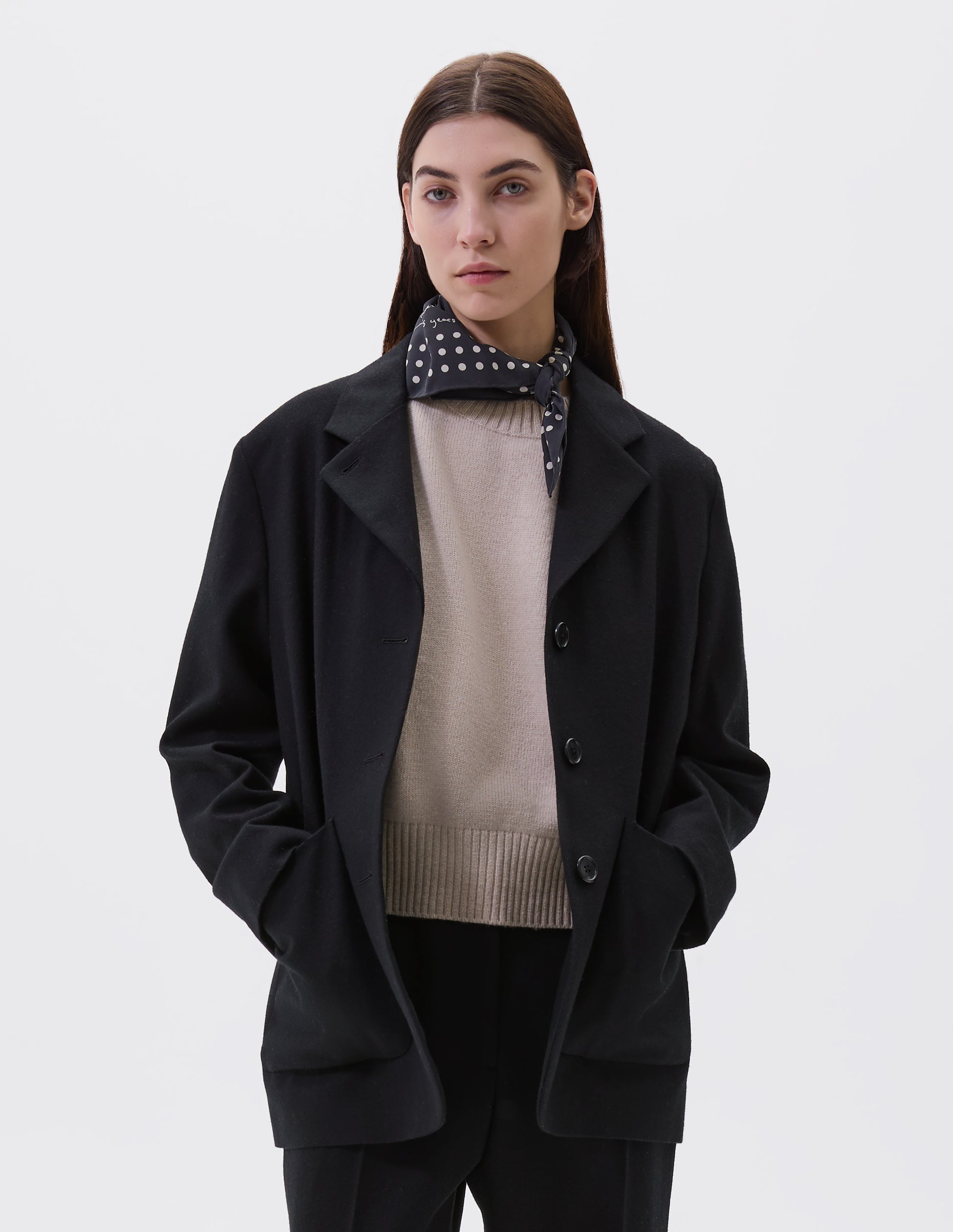 Cropped image, front view of model wearing black square neck scarf with white dots tied around neck. Styled with beige jumper, back blazer and matching black trousers.