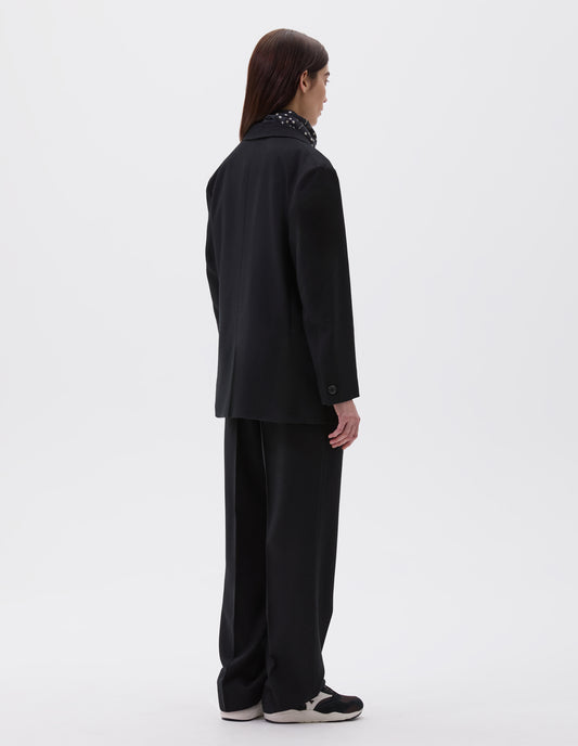 Full body image, back view of model wearing black relaxed blazer with horn buttons on cuffs and back vent. Styled with matching black trousers, polka dot neck scarf and black, brown and white trainers.