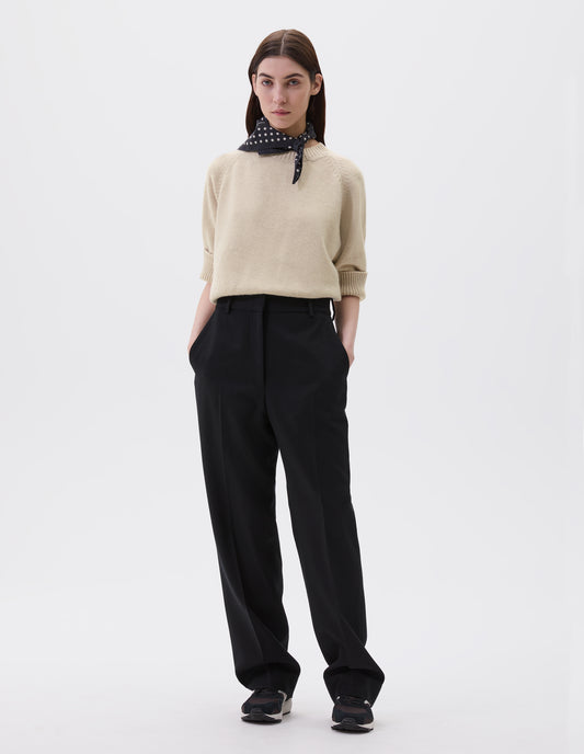 Full body image, front view of model wearing black long leg wool trousers, with belt loops and side pockets. Styled with beige short sleeve jumper, black and white polka dot scarf and black and brown trainers.
