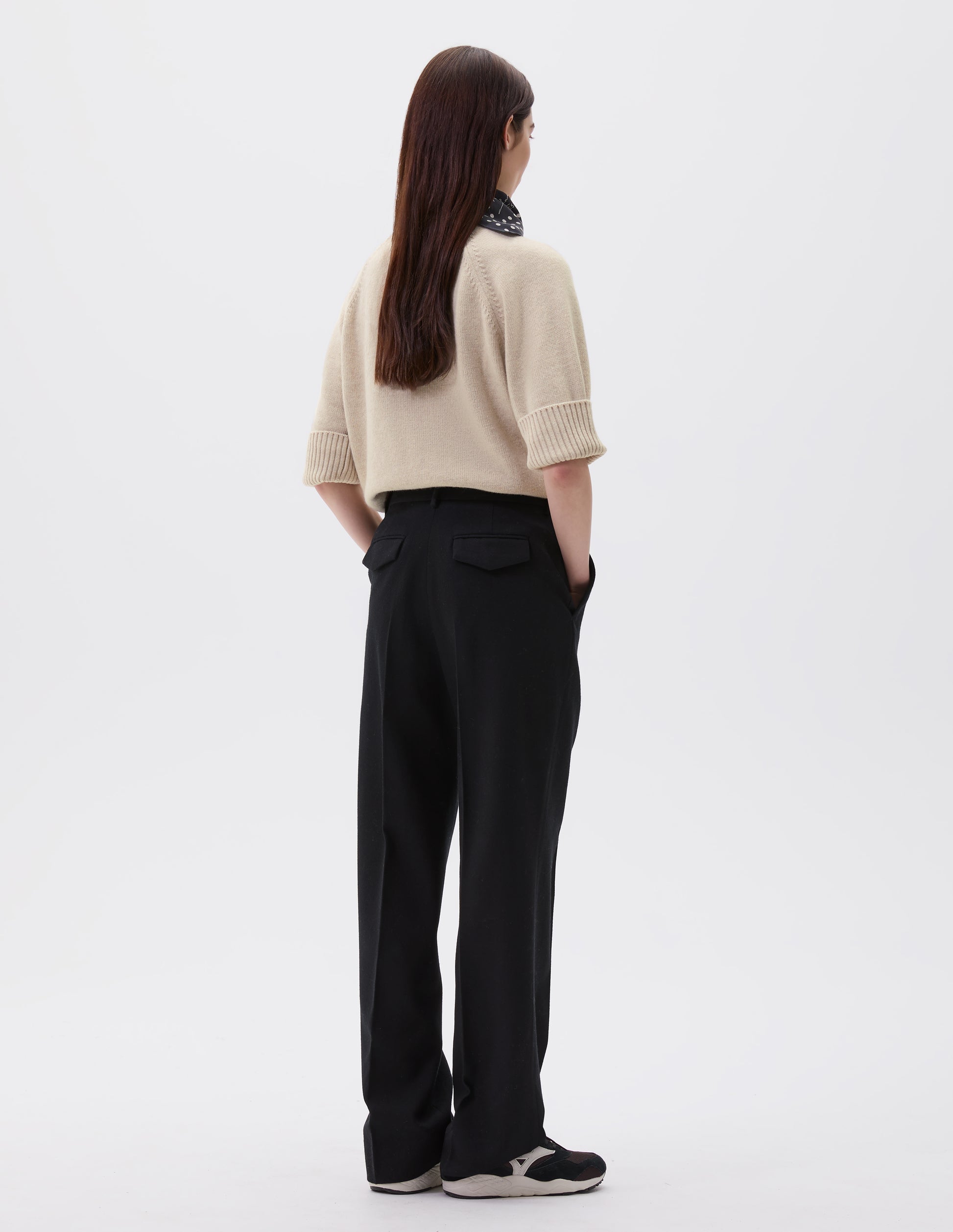 Full body image, back view of model wearing black long leg wool trousers, with belt loops, side pockets and concealed button fastening flap back pocket. Styled with beige short sleeve jumper, black and white polka dot scarf and black and brown trainers.