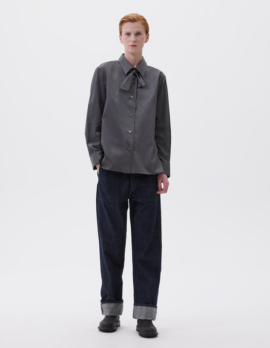 Full body image, front view of model wearing grey long sleeve shirt with wide stitch through placket, mother of pearl buttons, elongated back and removeable self fabric tie. Styled with indigo blue jeans and black leather boots.