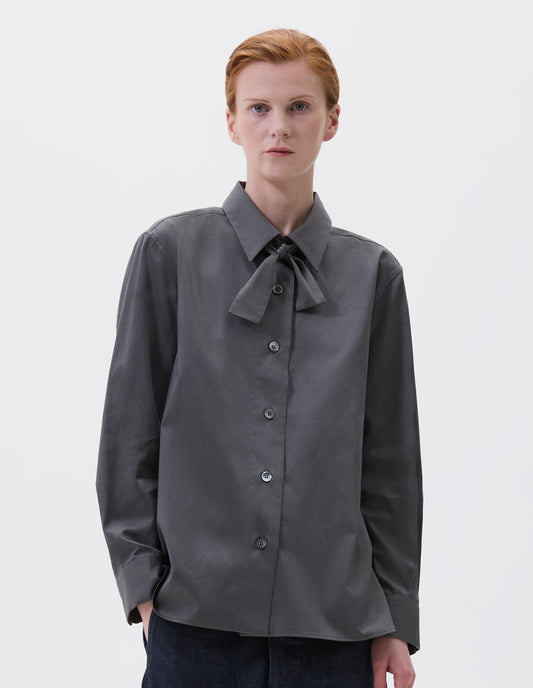 Cropped body image, front view of model wearing grey long sleeve shirt with wide stitch through placket, mother of pearl buttons, elongated back and removeable self fabric tie. Styled with indigo blue jeans.