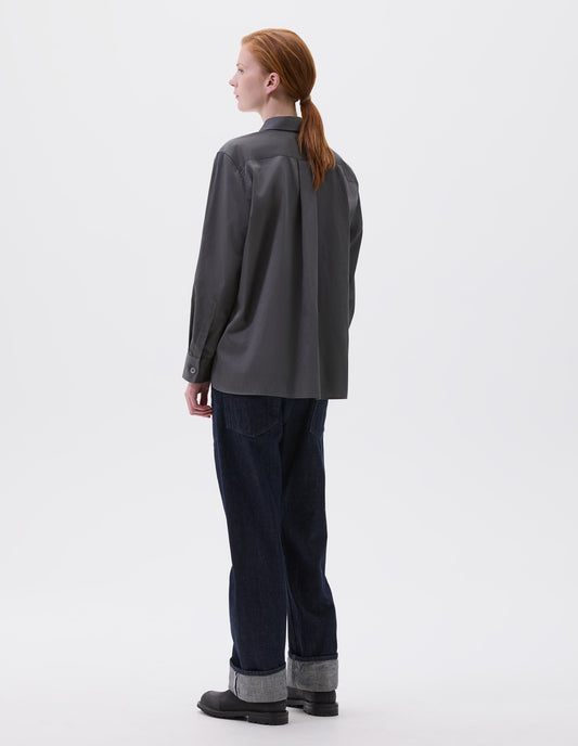 Full body image, back view of model wearing grey long sleeve shirt with with elongated back and mother of pearl button cuffs. Styled with indigo blue jeans and black leather boots.