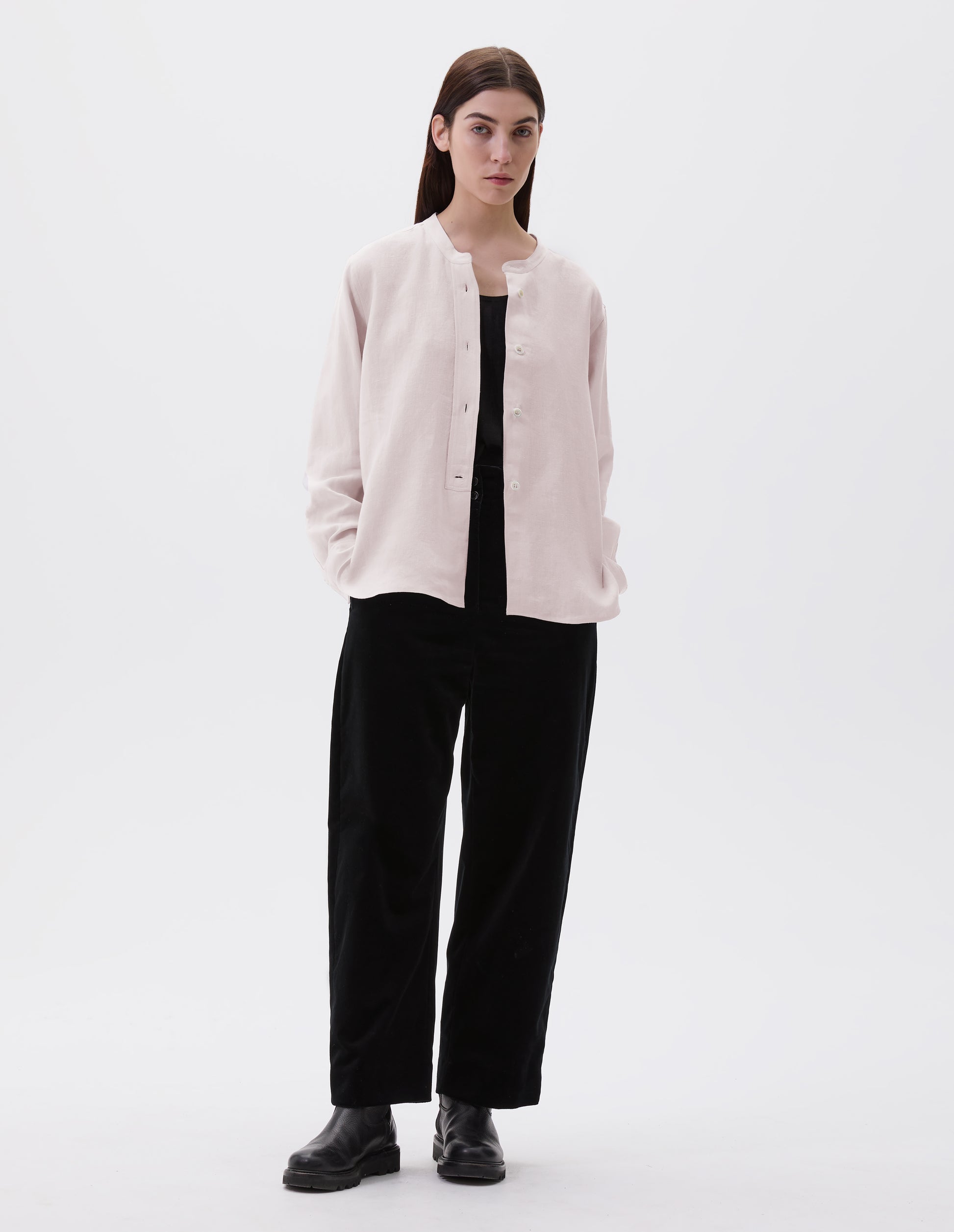 Full body image, front view of model wearing pinkish beige collarless shirt, with raised placket and mother of pearl buttons. Styled over black vest, black trousers and black leather boots. 