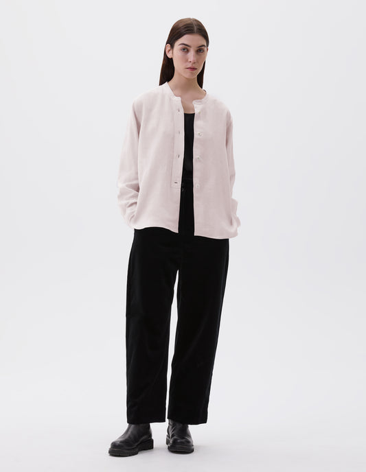 Full body image, front view of model wearing pinkish beige collarless shirt, with raised placket and mother of pearl buttons. Styled over black vest, black trousers and black leather boots. 