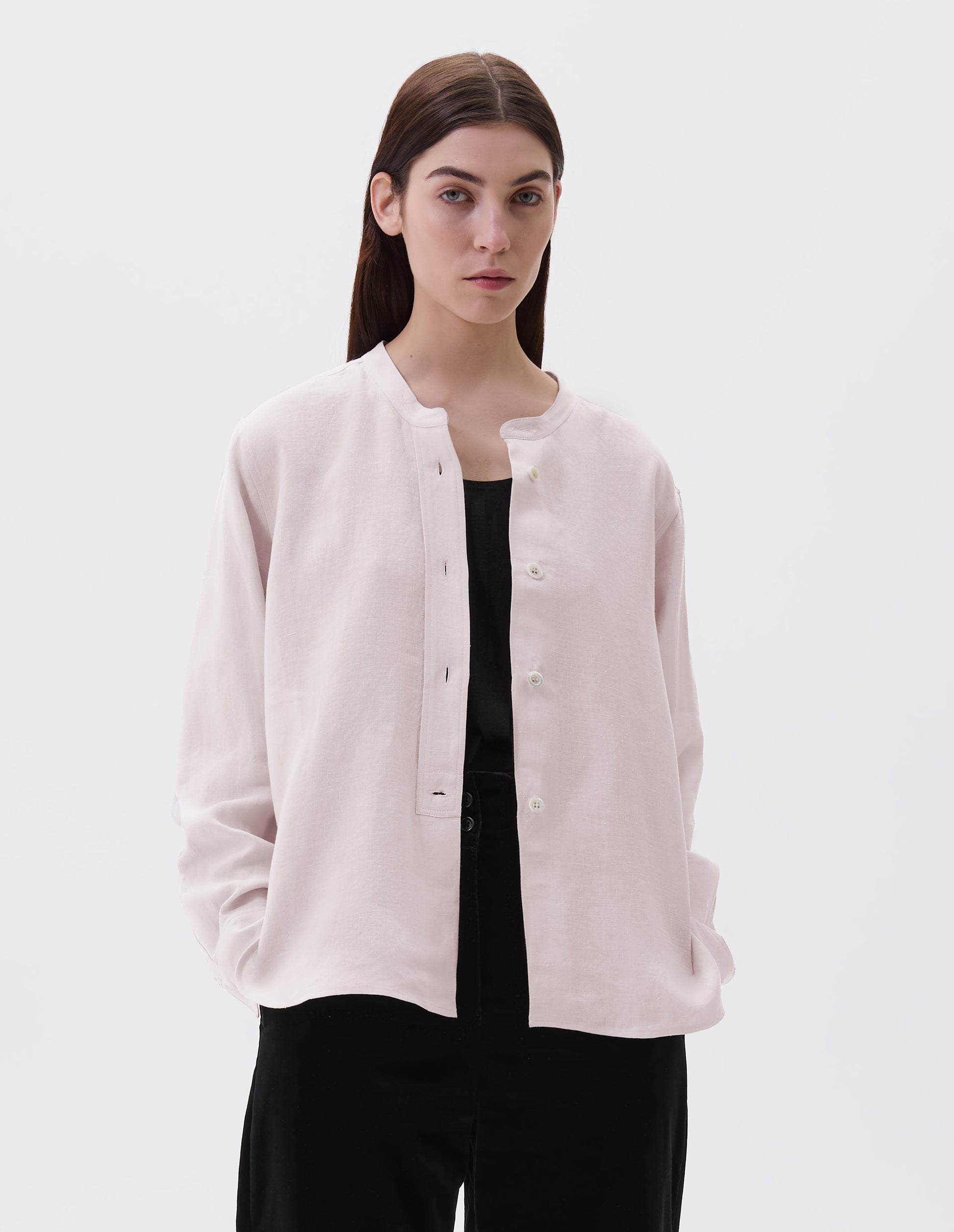 Cropped body image, front view of model wearing pinkish beige collarless shirt, with raised placket and mother of pearl buttons. Styled over black vest and black trousers.