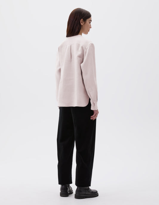 Full body image, back view of model wearing pinkish beige collarless shirt, with mother of pearl button cuffs, box stitch detailing on back yoke and elongated hem. Styled over black vest, black trousers and black leather boots. 