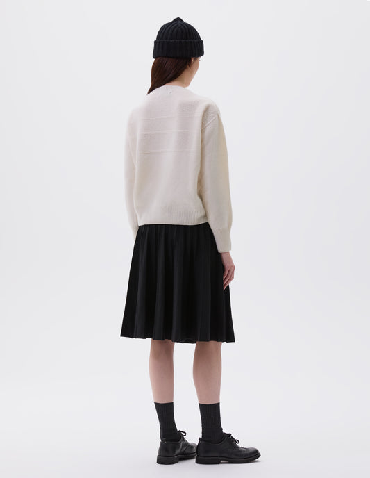 Full body image, back view of model wearing ecru crew neck jumper with textured panelling on shoulders and back and contrasting rib on neck, cuffs and hem. Styled with black rib beanie, black knitted skirt, black socks and black leather brogues.