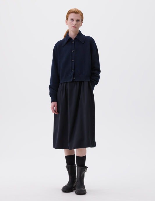 Full body image, front view of model wearing dark blue boxy cropped cardigan with collar, horn buttons and contrast rib on collar, cuffs and hem. Styled over dark blue dress and black socks with black leather biker boots.
