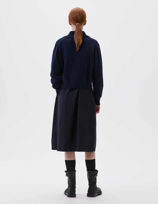 Full body image, back view of model wearing dark blue boxy cropped cardigan with collar, contrast rib on collar, cuffs and hem. Styled over dark blue dress and black socks with black leather biker boots.