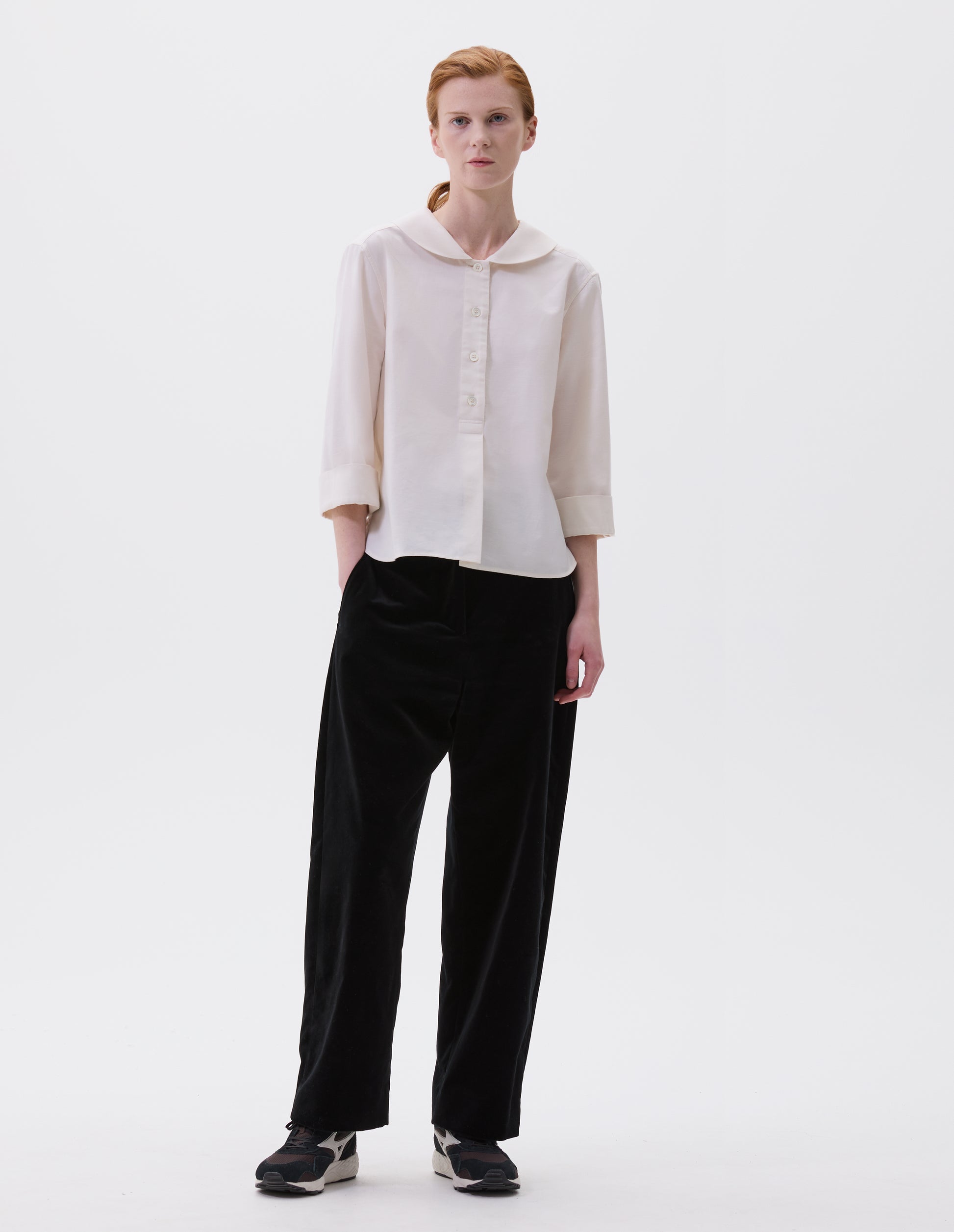 Full body image, front view of model wearing off white petal collar shirt with three quarter length sleeves, stitch through half placket, mother of pearl buttons and turned back cuff details. Styled with black velvet trousers and black and brown trainers.