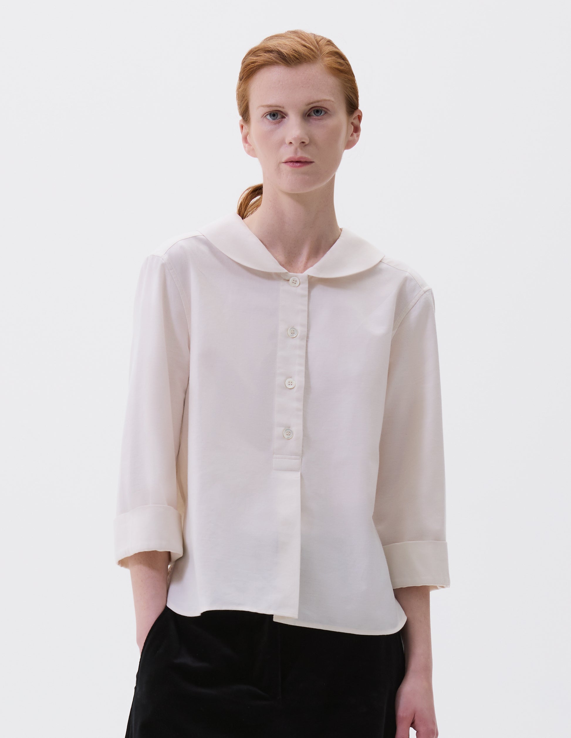 Cropped body image, front view of model wearing off white petal collar shirt with three quarter length sleeves, stitch through half placket, mother of pearl buttons and turned back cuff details. Styled with black velvet trousers.