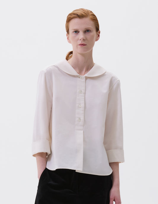 Cropped body image, front view of model wearing off white petal collar shirt with three quarter length sleeves, stitch through half placket, mother of pearl buttons and turned back cuff details. Styled with black velvet trousers.