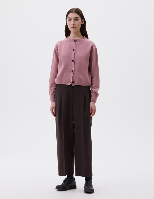 Full body image, front view of model wearing pink cardigan with contrasting rib neckline, hem and cuffs and dark horn buttons. Styled with brown trousers, black socks and black leather brogues.