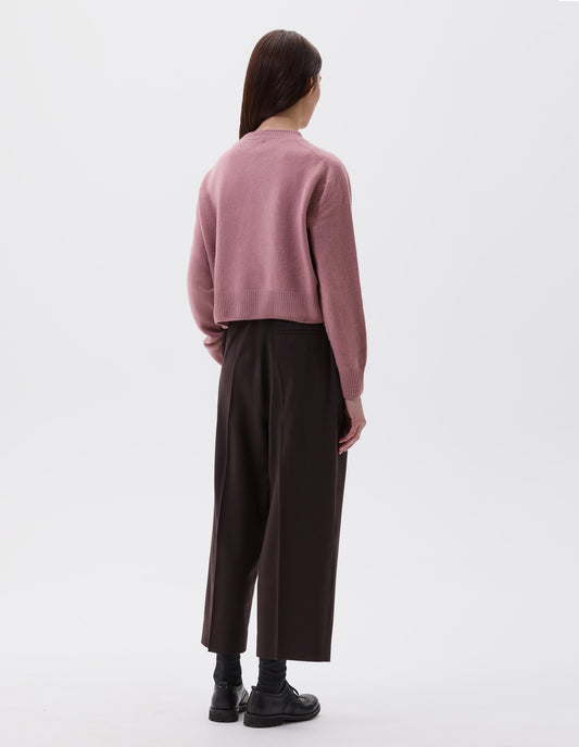 Full body image, back view of model wearing relaxed dark brown pleated cropped trousers with welt back pocket. Styled with pink boxy cardigan, black socks and black leather brogues.
