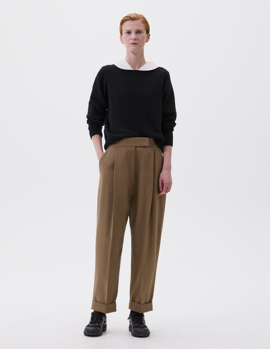 Full body image, front view of model wearing relaxed dark beige pleated and tapered trousers with hook and bar fastening, side pockets and cuffed hems. Styled with black jumper over white shirt and black trainers. 