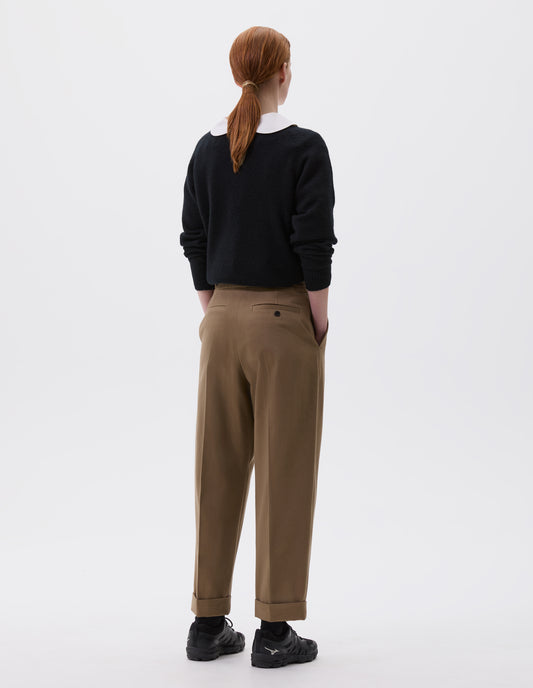 Full body image, back view of model wearing relaxed dark beige pleated and tapered trousers with side pockets, welt back pocket and button-through welt back pocket, cuffed hems. Styled with black jumper over white shirt and black trainers. 