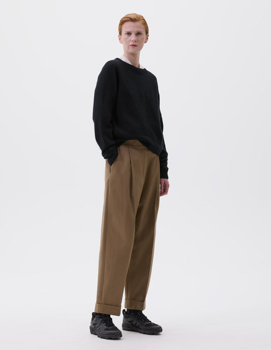 Full body image, front view of model wearing black long sleeve cashmere jumper, with raglan sleeve and contrasting rib on neck, cuffs and hem. Styled over white collared shirt, with brown pleated trousers and black trainers.