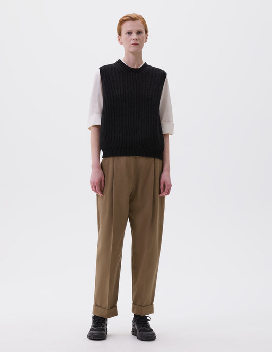 Full body image, front view of model wearing black sleeveless merino wool slipover, with contrast rib on neck, armhole and hem. Styled with beige pleated trousers, white shirt and black trainers.