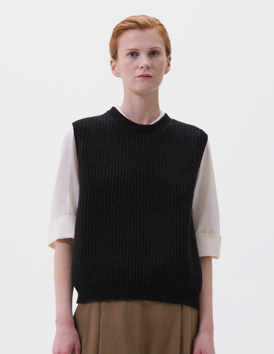 Cropped body image, front view of model wearing black sleeveless merino wool slipover, with contrast rib on neck, armhole and hem. Styled with beige pleated trousers and white shirt.