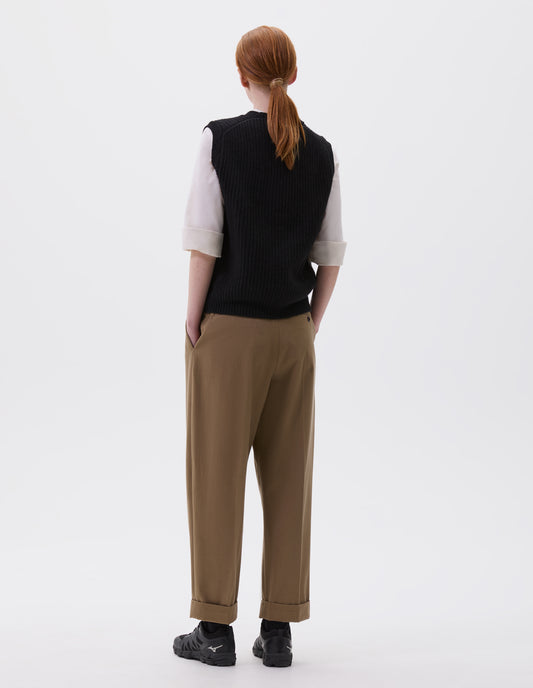 Full body image, back view of model wearing black sleeveless merino wool slipover, with contrast rib on neck, armhole and hem. Styled with beige pleated trousers, white shirt and black trainers.