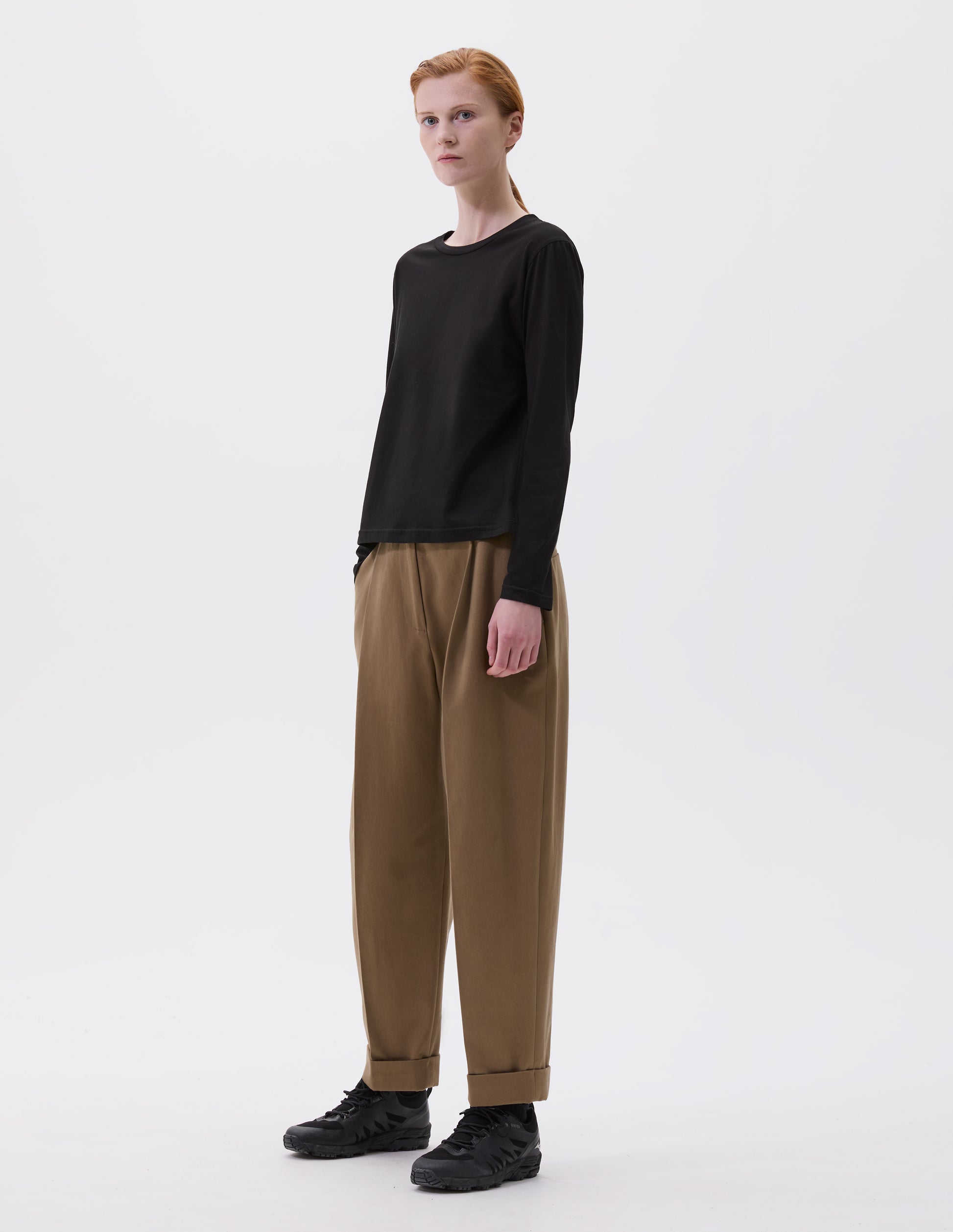 Full body image, front view of model wearing black cotton wide neck long sleeve with boxy cut. Styled with brown trousers and black trainers.