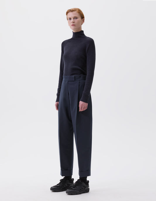Full body image, front view of model wearing dark grey fine knit merino wool jumper, with high neck, raglan sleeve and cast off edging on neck, cuffs and hem. Styled navy blue pinstripe pleated trousers, black socks and black trainers.