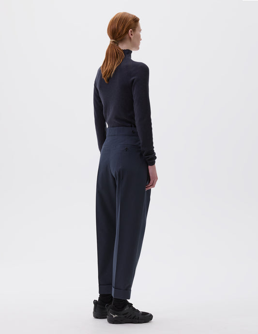Full body image, back view of model wearing relaxed dark blue tapered trousers with grey stripes, pleats, cuffed hems and button-through welt back pocket, styled with dark grey turtleneck, black socks and black trainers.