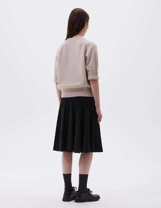Full body image, back view of model wearing black relaxed knee length knitted skirt with pleats. Styled with beige jumper, black socks and black leather brogues.