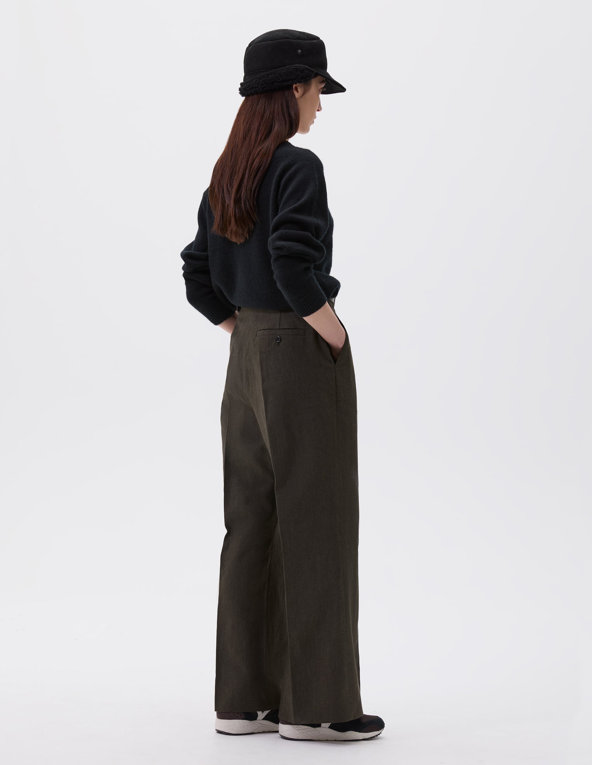 Full body image, back view of model wearing relaxed brown trousers with straight leg and welt button-through back pocket. Styled with black shearling cap, black jumper and black, brown and white trainers.