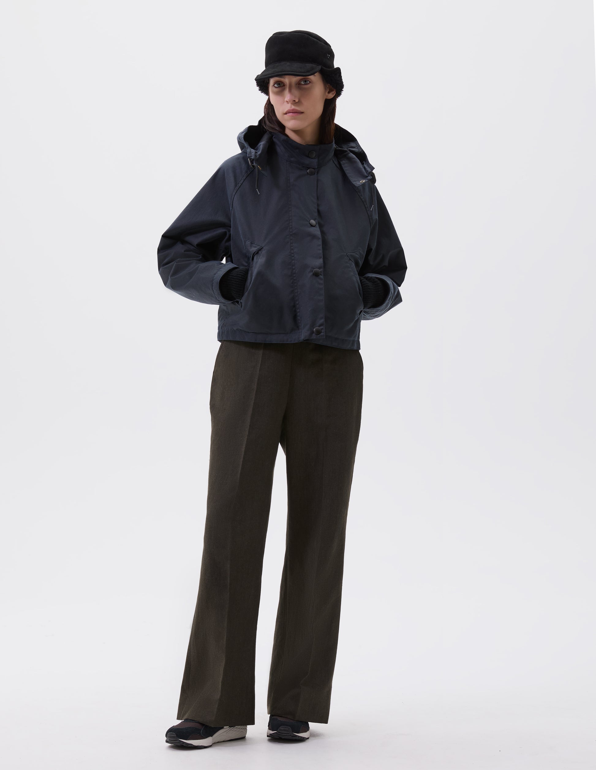 Full body image, front view of model wearing boxy dark blue jacket with stand collar and adjustable hood, raglan sleeve, press stud storm flap and wide storm welt side pockets. Styled with black shearling cap, brown trousers and black, brown and white trainers.