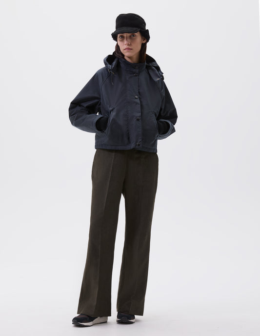Full body image, front view of model wearing boxy dark blue jacket with stand collar and adjustable hood, raglan sleeve, press stud storm flap and wide storm welt side pockets. Styled with black shearling cap, brown trousers and black, brown and white trainers.