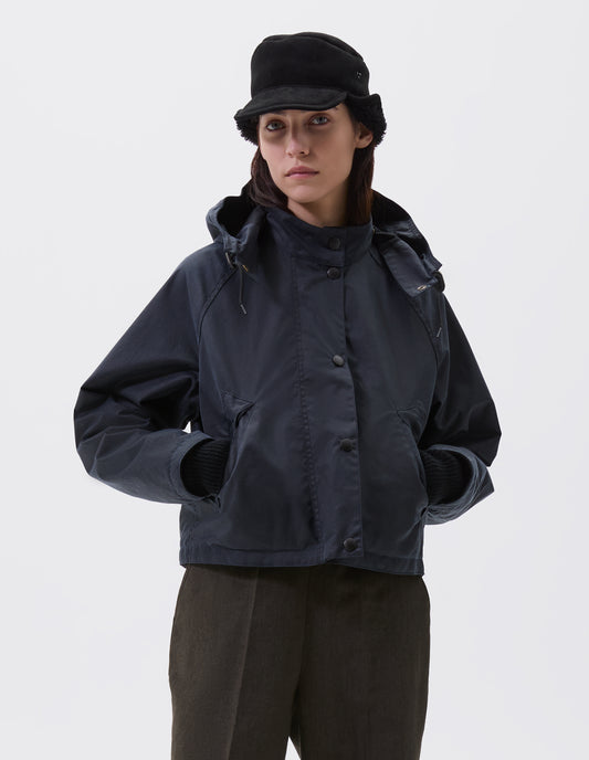Cropped image, front view of model wearing boxy dark blue jacket with stand collar and adjustable hood, raglan sleeve, press stud storm flap and wide storm welt side pockets. Styled with black shearling cap and brown trousers.