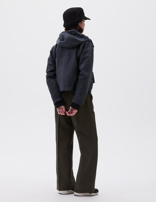 Full body image, back view of model wearing boxy dark blue jacket with raglan sleeve and hood with wire adjustable peak. Styled with black shearling cap, brown trousers and black, brown and white trainers.