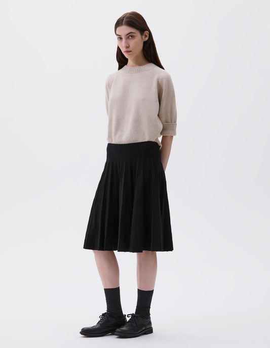 Full body image, front view of model wearing black relaxed knee length knitted skirt with pleats. Styled with beige jumper, black socks and black leather brogues.