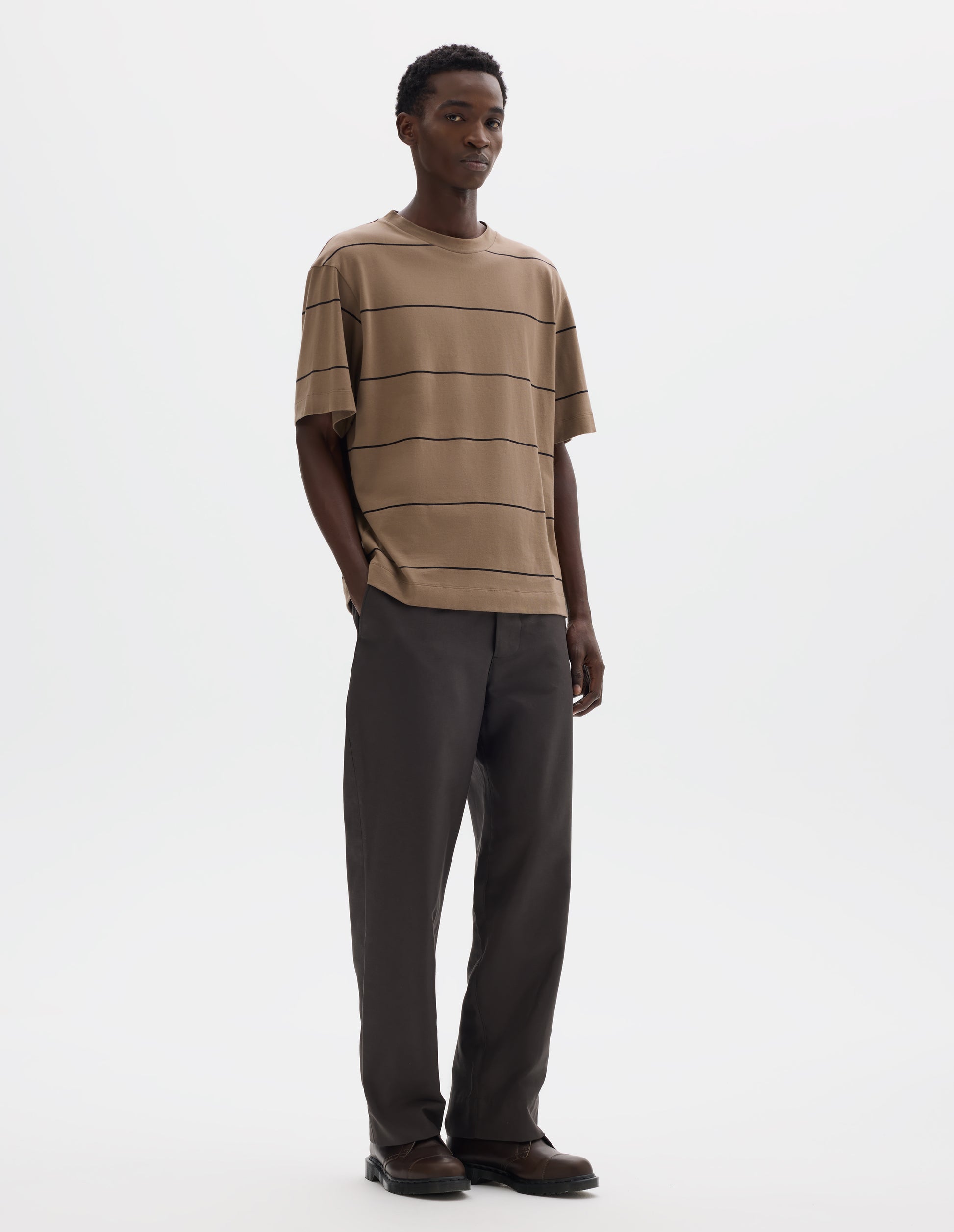 Full body image, front view of model wearing  relaxed beige jersey t-shirt with black stripes, boxy cut and with crew neck. Styled with grey trousers and brown leather shoes.