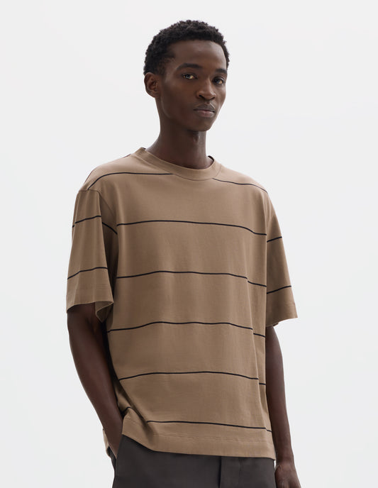 Cropped body image, front view of model wearing relaxed beige jersey t-shirt with black stripes, boxy cut and with crew neck. Styled with grey trousers.