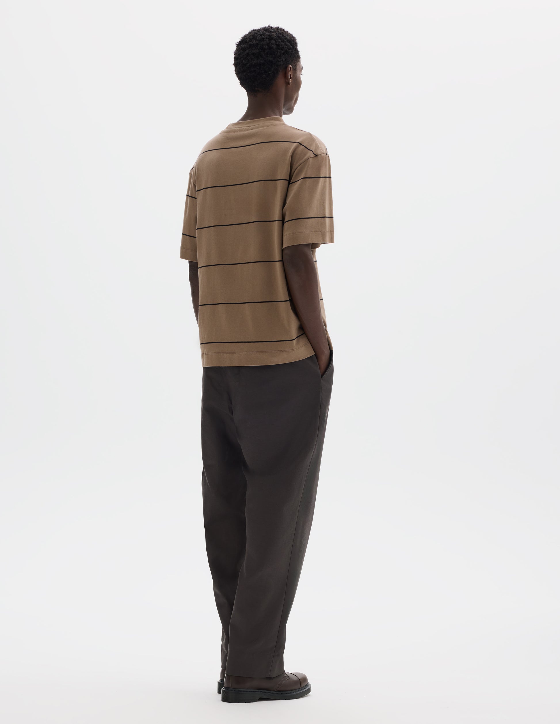 Full body image, back view of model wearing relaxed beige jersey t-shirt with black stripes, boxy cut and with crew neck. Styled with grey trousers and brown leather shoes.