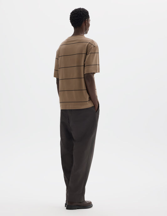 Full body image, back view of model wearing relaxed beige jersey t-shirt with black stripes, boxy cut and with crew neck. Styled with grey trousers and brown leather shoes.