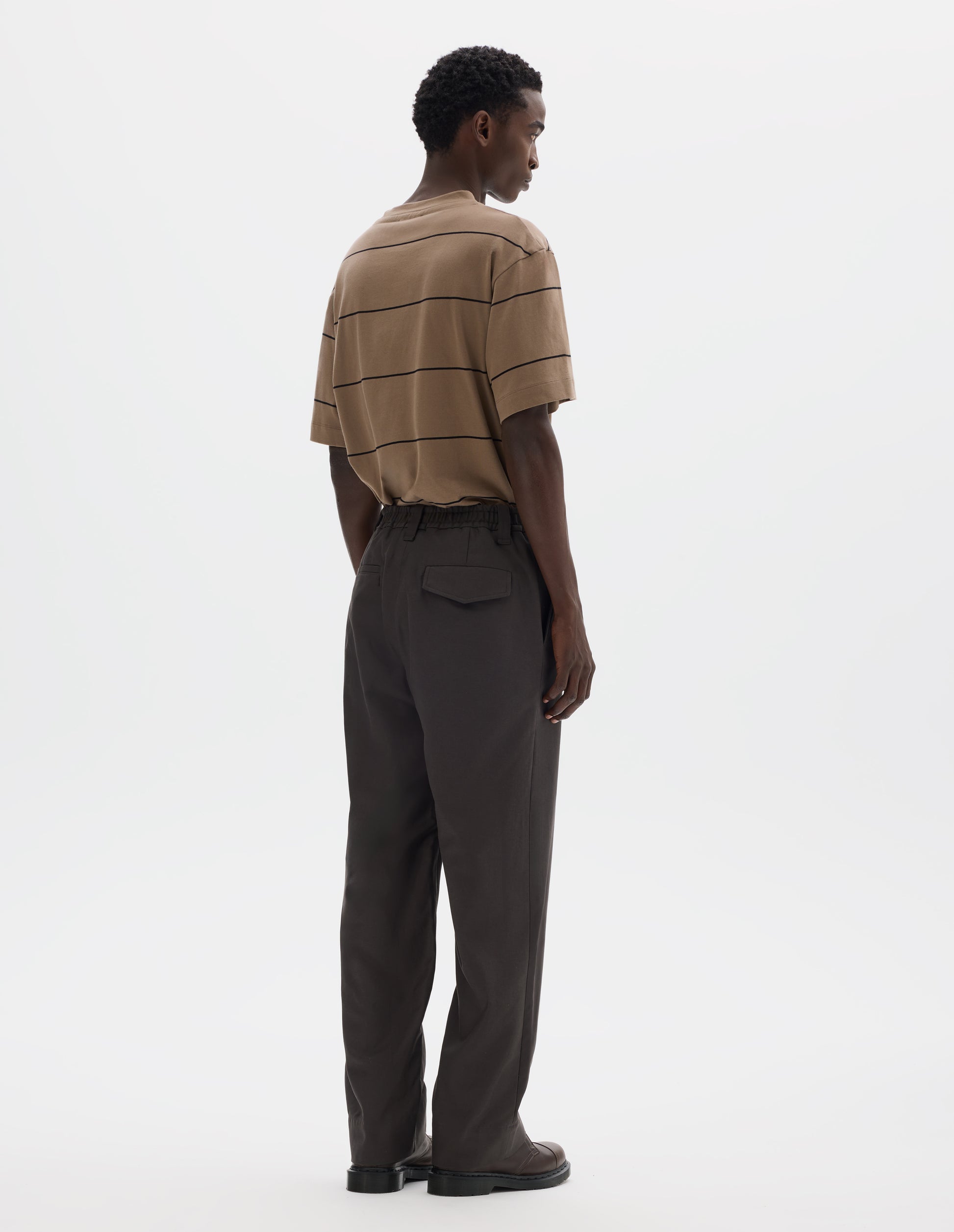 Full body image, back view of model wearing relaxed grey cotton trousers with half-elasticated waistband, belt loops, slant side pockets, and welt and flap back pocket with concealed horn button fastening. Styled with beige striped t-shirt and brown leather shoes.