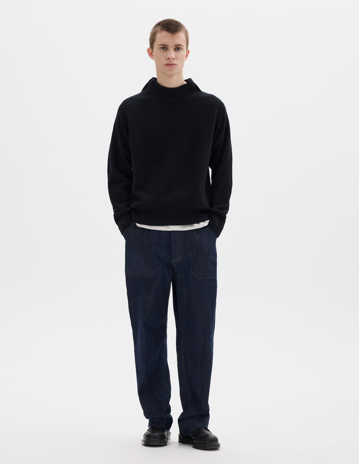 Full body image, front view of model wearing black fully fashioned seamless wool jumper with rib mock neck, set-in sleeves, and ribbed cuffs and hem. Styled over white t-shirt, and with dark blue jeans and black leather shoes.