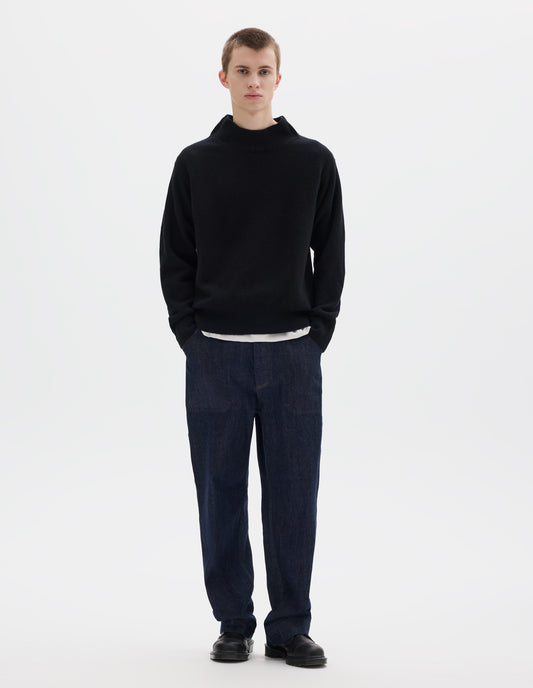 Full body image, front view of model wearing black fully fashioned seamless wool jumper with rib mock neck, set-in sleeves, and ribbed cuffs and hem. Styled over white t-shirt, and with dark blue jeans and black leather shoes.