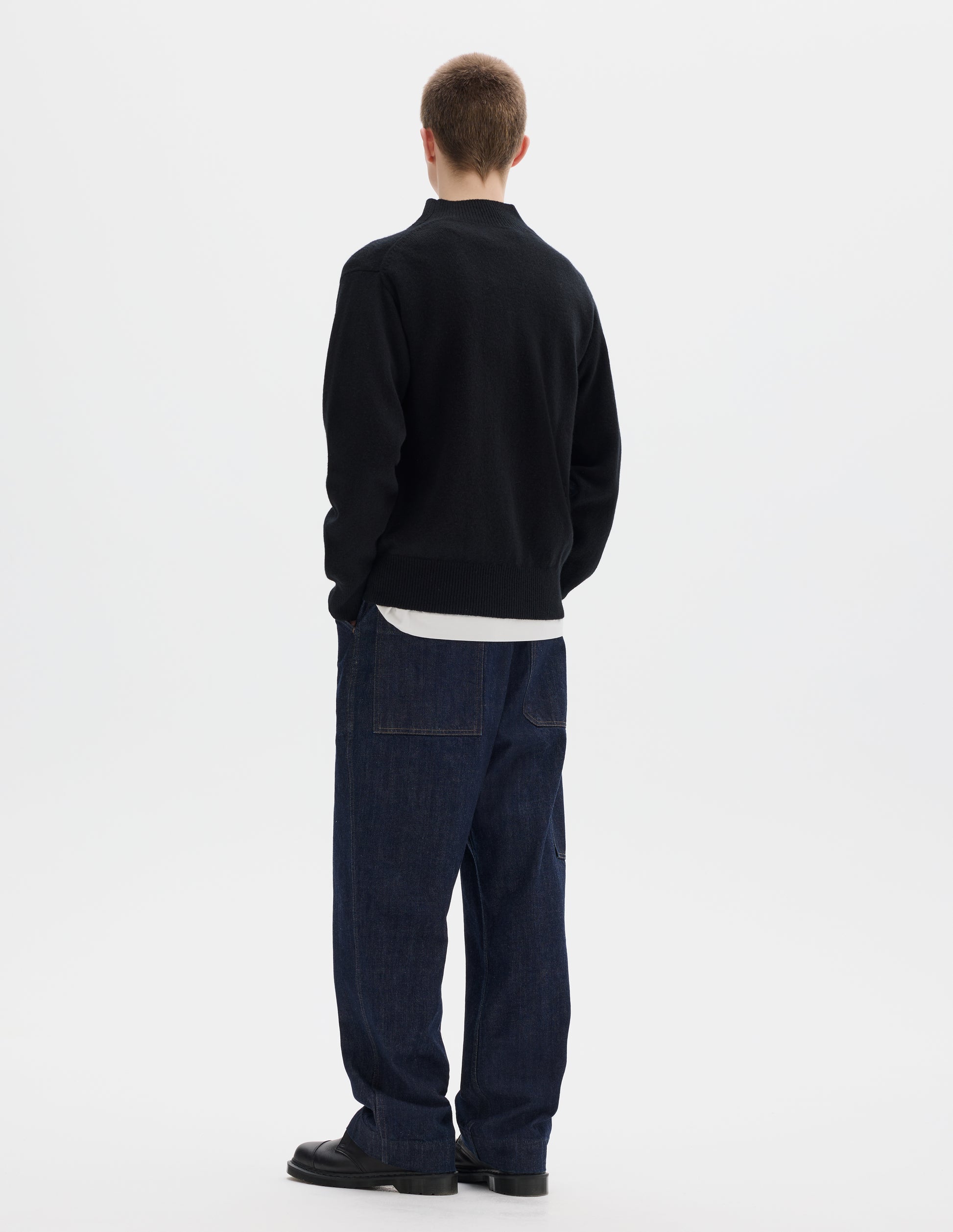 Full body image, back view of model wearing black fully fashioned seamless wool jumper with rib mock neck, set-in sleeves, and ribbed cuffs and hem. Styled over white t-shirt, and with dark blue jeans and black leather shoes.