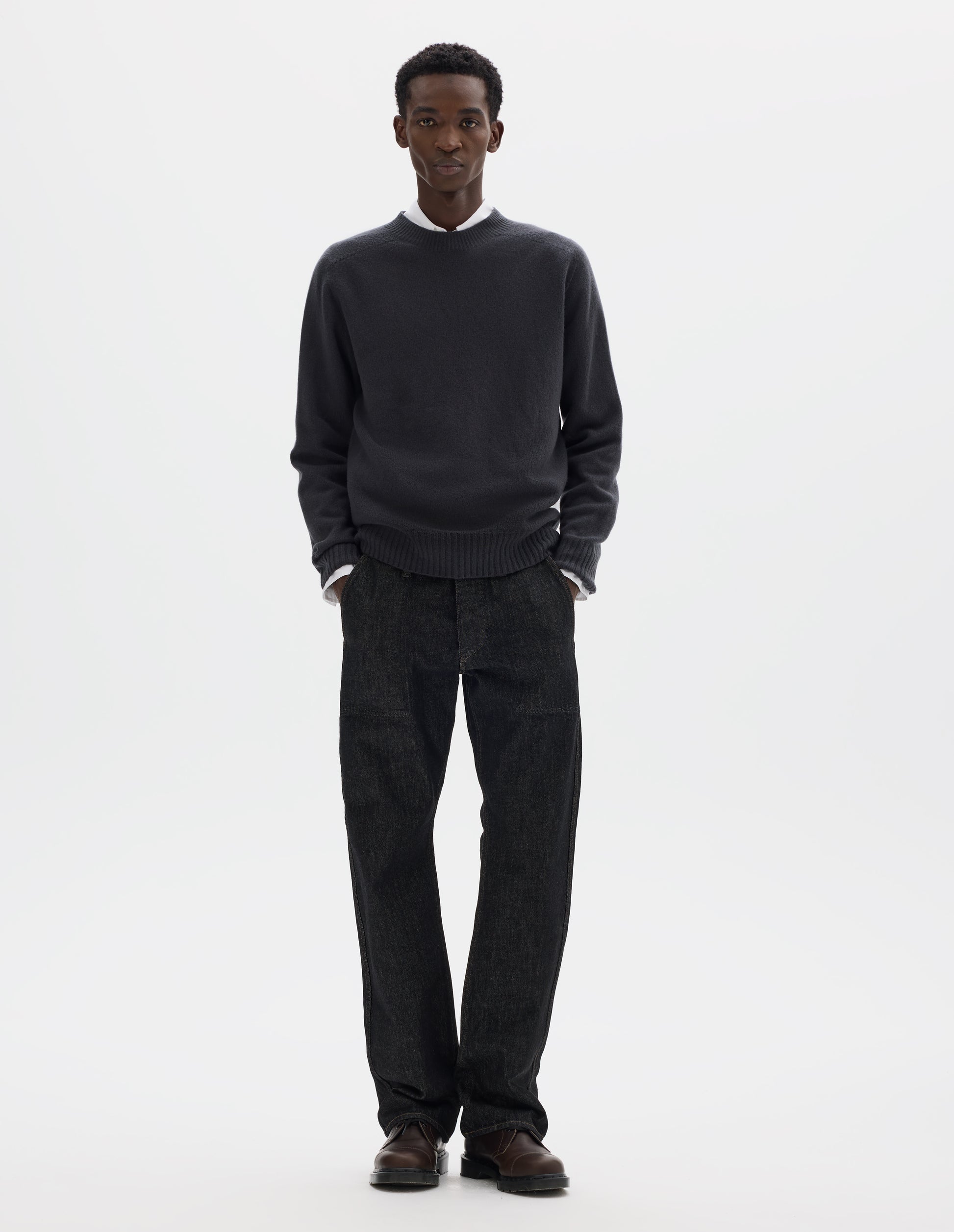 Full body image, front view of model wearing dark grey merino cashmere seamless jumper with ribbed crew neck, cuffs and hem. Styled over white shirt, and with black jeans and brown leather shoes.
