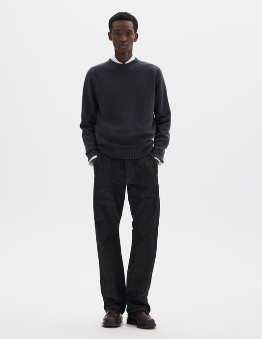 Full body image, front view of model wearing dark grey merino cashmere seamless jumper with ribbed crew neck, cuffs and hem. Styled over white shirt, and with black jeans and brown leather shoes.
