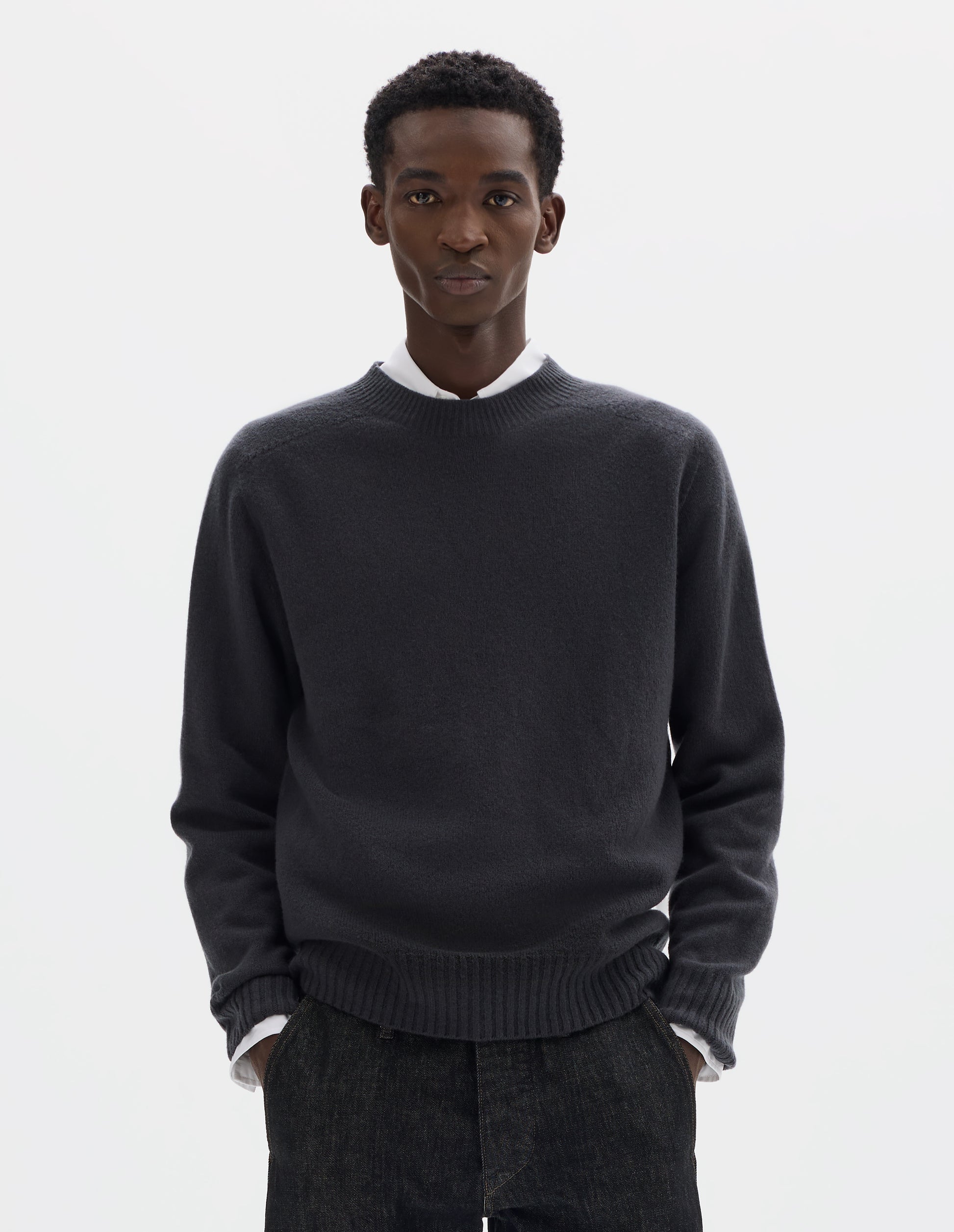 Cropped body image, front view of model wearing dark grey merino cashmere seamless jumper with ribbed crew neck, cuffs and hem. Styled over white shirt, and with black jeans.